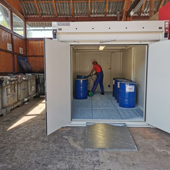 Eine Person schiebt einen Trolley mit blauen Fässern in einem offenen Lagercontainer mit Metallboden.
