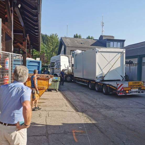Ein Mann beobachtet, wie ein Lkw einen großen Container neben einem Gebäude mit einem Müllcontainer entlädt. Zwei Arbeiter sind in der Nähe, einer steht neben dem Müllcontainer. Bäume und ein klarer Himmel sind im Hintergrund.
