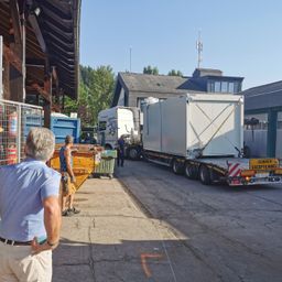 Ein Mann beobachtet, wie ein Lkw einen großen Container neben einem Gebäude mit einem Müllcontainer entlädt. Zwei Arbeiter sind in der Nähe, einer steht neben dem Müllcontainer. Bäume und ein klarer Himmel sind im Hintergrund.