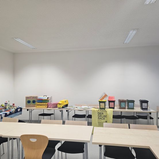Ein leerer Klassenraum mit Tischen und Stühlen, einem langen Schreibtisch mit Behältern und einem gelben Beutel. Auf dem Schreibtisch befinden sich verschiedene Gegenstände, darunter Bücher, eine Schachtel und ein gelber Spielzeugwagen. Der Raum hat weiße Wände und eine weiße Decke mit einem Netzmuster.