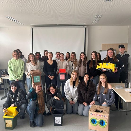 Eine Gruppe von Personen versammelt sich in einem Klassenzimmer und posiert für ein Foto mit verschiedenen Behältern. Jede Person hält einen Behälter, und einige lächeln. Das Klassenzimmer hat weiße Wände und Deckenleuchten.