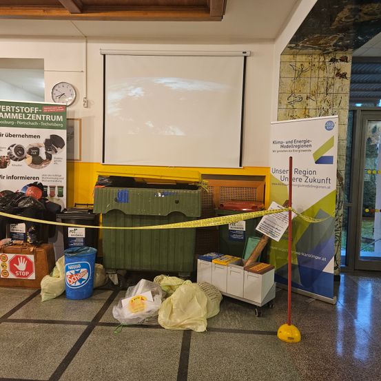 Ein Raum mit gelbem Absperrband auf dem Boden, mehreren Mülleimern, einem blauen Eimer und einem Banner mit dem Text Unsere Region Unsere Zukunft auf der rechten Seite.