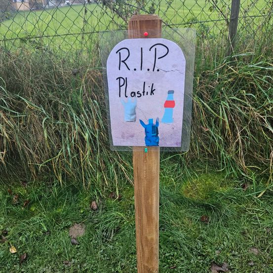 Ein Holzpfosten mit einem Schild, auf dem in Kreide R.I.P. Plastik steht, mit einer Zeichnung einer Flasche und eines Beutels darauf.