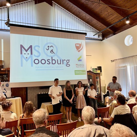 Eine Gruppe junger Menschen steht auf der Bühne mit einer Projektionsleinwand hinter ihnen, die eine Präsentation über Moosburg zeigt. Ein Publikum sitzt vor ihnen.