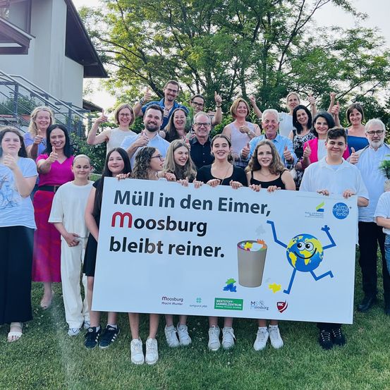 Eine Gruppe von Menschen in Freizeitkleidung posiert für ein Foto auf einem Rasen. Sie halten ein Banner mit einem Cartoon-Charakter und dem Text 'Mull in den Eimer, Moosburg bleibt reiner.' Im Hintergrund befindet sich ein Haus und einige Bäume.