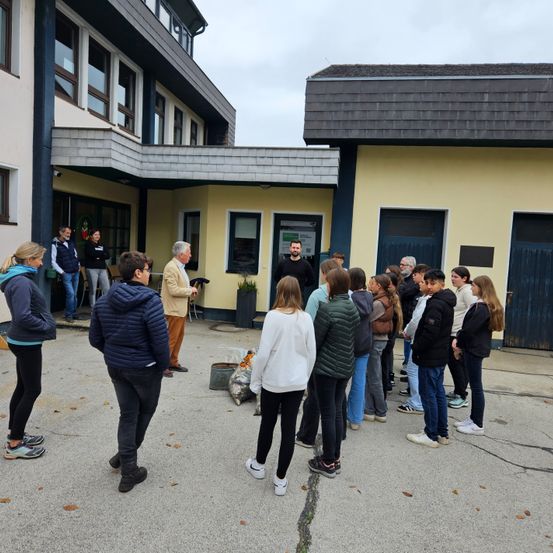 Eine Gruppe von Menschen, darunter ein sprechender Mann, steht vor einem gelben Gebäude mit vielen Fenstern. Einige tragen Jacken und einige haben Turnschuhe an.