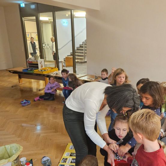 Ein Raum mit Kindern, die um eine Frau herum sitzen und stehen. Einige Kinder betrachten eine Geschenkbox, die die Frau öffnet.