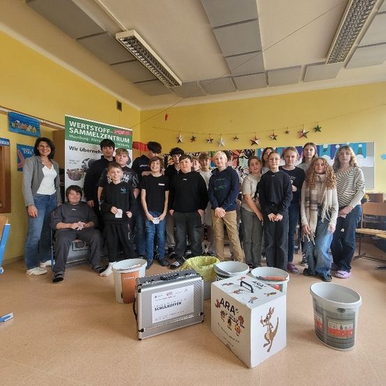 Eine Gruppe von Kindern und Erwachsenen posiert für ein Foto in einem Klassenzimmer mit einem Banner, Eimern und einer Aufbewahrungsbox. Sie tragen alle Freizeitkleidung.