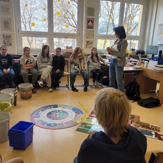 Eine Frau steht vor einem Klassenzimmer, hält ein Papier in der Hand, während eine Gruppe von Schülern auf Stühlen sitzt und zuhört. Das Klassenzimmer hat Fenster mit Bäumen draußen und einen Schreibtisch mit Computermonitor. Es gibt Eimer, einen Mülleimer und einen Teppich auf dem Boden.