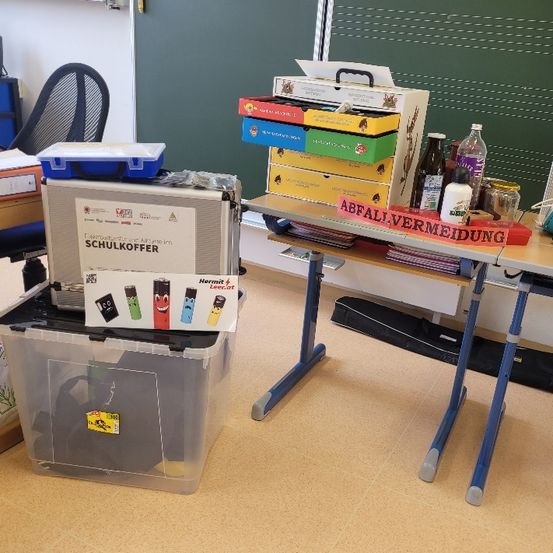 Ein Klassenzimmer mit einem Klapptisch, gefüllt mit Kisten und Behältern. Auf dem Tisch stehen Bücher, Flaschen und ein Schild mit der Aufschrift 'Abfallvermeidung'. Daneben befinden sich Lagerboxen und eine schwarze Tasche. Hinter dem Tisch steht eine grüne Tafel.