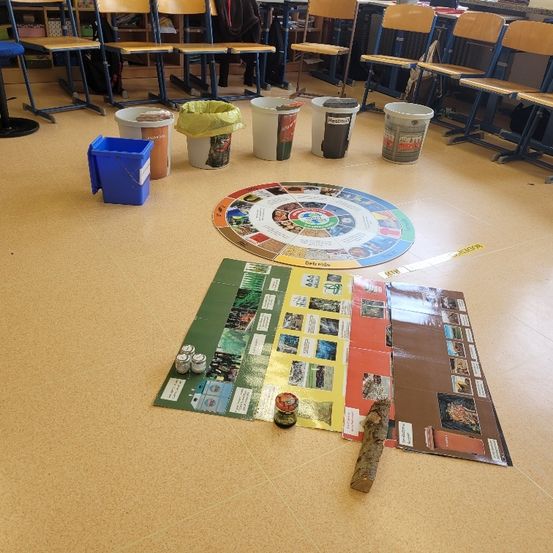 Ein Innenraum mit einem Recycling-Spielbrett, fünf Eimern und einem blauen Behälter. Das Spielbrett ist kreisförmig, und es gibt verschiedene Karten auf dem Boden. Stühle sind im Hintergrund aufgereiht.