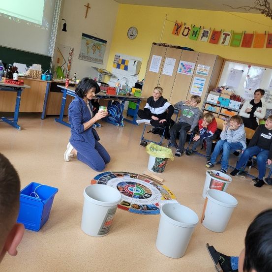 In einem Klassenzimmer kniet eine Frau mit einem Becher in der Hand, umgeben von Kindern, die im Kreis sitzen, mit Eimern und einer runden Tafel auf dem Boden.