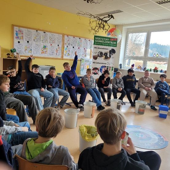 Eine Gruppe von Schülern und Erwachsenen sitzt in einem Klassenzimmer mit Recycling-Behältern, Postern an der Wand und einem Banner mit der Aufschrift 'Wertstoffsammlung Sammelzentrum.' Sie scheinen an einer Aktivität beteiligt zu sein.