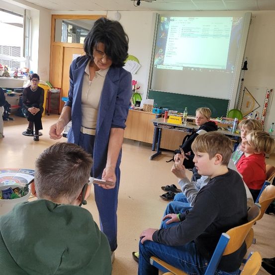 Eine Frau in einem blauen Blazer steht in einem Klassenzimmer und zeigt Schülern ein kleines Objekt. Sie sitzen in Stühlen und beobachten aufmerksam. Eine Projektionsleinwand und eine Tafel sind im Hintergrund.