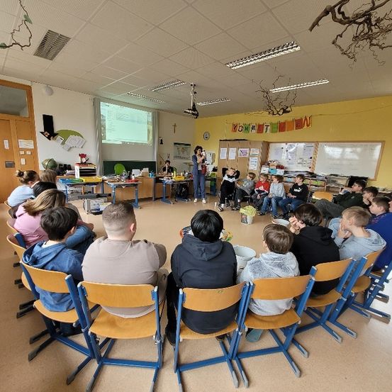 Ein Klassenzimmer mit Schülern und einer Lehrerin im Kreis, mit einer Projektionsleinwand und verschiedenen Dekorationen an den Wänden.