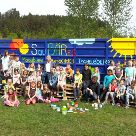 Eine Gruppe von Kindern und Erwachsenen posiert für ein Foto vor einem bunt bemalten Container. Bäume sind im Hintergrund zu sehen.