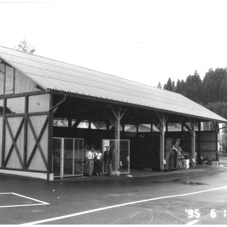 Ein Schwarz-Weiß-Foto eines Gebäudes mit geneigtem Dach, Holzbalken und einem Zaun. Drei Personen stehen am Eingang, und in der Nähe ist ein Fahrrad geparkt.