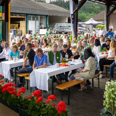 Eine Gruppe von Menschen sitzt an Tischen im Freien, umgeben von roten Blumen, mit Gebäuden und Bäumen im Hintergrund.