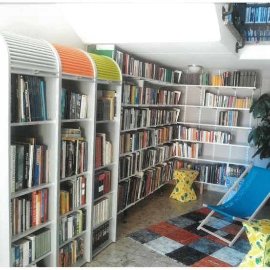 Eine kleine Bibliothek mit weißen Regalen voller Bücher. Bunte Stühle und ein Teppich sind im Raum. Die Regale sind ordentlich organisiert und die Bücher sind in Ordnung angeordnet.