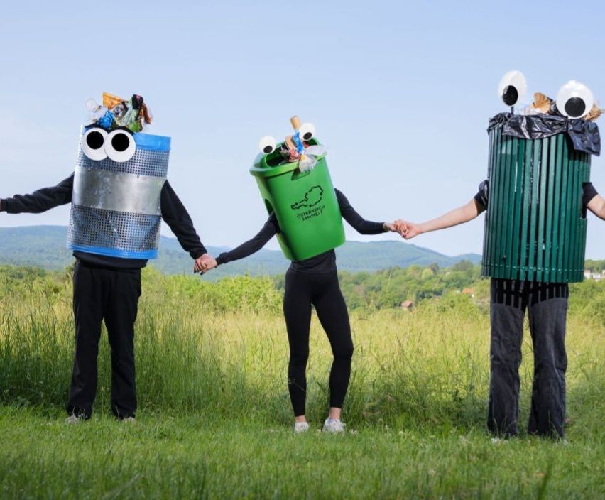 Drei Menschen in Mülleimer-Kostümen stehen in einem Grasfeld und halten Händchen. Einer trägt einen blauen Behälter, ein anderer einen grünen und der dritte einen Müllsack. Berge sind im Hintergrund.