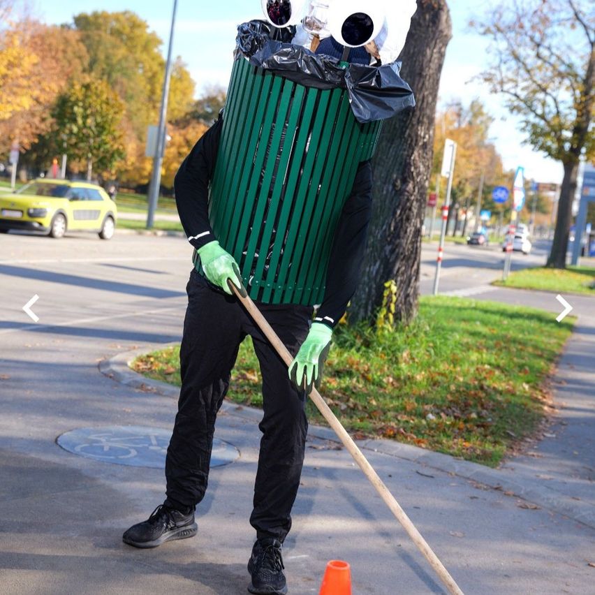 Eine Person, die als Mülltonne verkleidet ist, hält eine Kehrschaufel auf einem Bürgersteig. Die Mülltonne ist mit Plastiktüten gefüllt. Im Hintergrund sind Autos zu sehen.