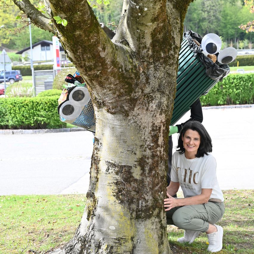 Eine Frau kniet neben einem Baum mit einem Mülleimer mit Kulleraugen. Sie hält den Baumstamm und lächelt. Eine Person in einer grünen Mülleimer-Verkleidung steht hinter dem Baum.