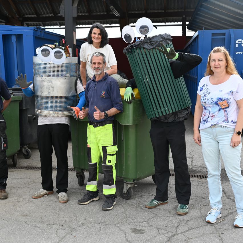 Fünf Personen, darunter zwei in Kostümen, stehen neben Mülltonnen im Freien und lächeln für das Foto.
