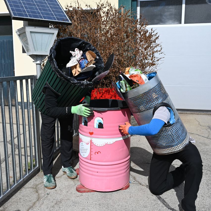 Zwei Menschen in Kostümen leeren Mülleimer in einen großen rosafarbenen Fass. Über den Mülleimern ist ein Solarpanel montiert.