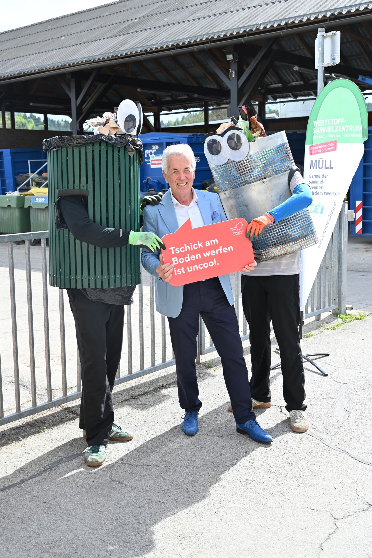 Ein Mann in einem blauen Anzug, der ein Schild hält, flankiert von zwei Personen in Mülleimerkostümen, eine mit einem Mülleimerhelm, die andere mit einem Mülleimerkostüm mit Augen.