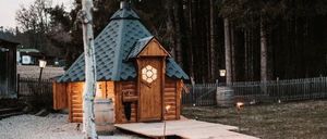 Eine kleine Holzhütte mit schwarzem Dach und einem hölzernen Gehweg, der zu ihr führt. Die Hütte ist beleuchtet und von einem Zaun umgeben. Bäume befinden sich im Hintergrund.