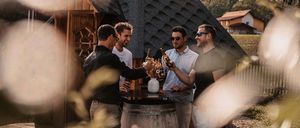 Vier Männer stehen um einen Fass herum, stoßen mit Flaschen an, vor einem Haus mit einem schiefergedeckten Dach, in einem ländlichen Gebiet.