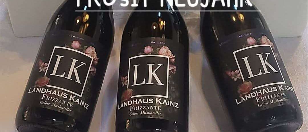Drei Flaschen Landhaus Kainz Frizzante Wein sind mit roten Pilzen im Hintergrund ausgestellt. Das Etikett sagt Prosit Neujahr. Der Text darunter sagt Ab 2. Januar sind wir wieder fur euch da.