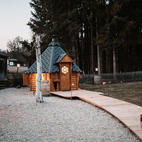 Eine Holzhütte mit grauem Dach steht auf einem Kiesweg in einem bewaldeten Gebiet, mit Bäumen und einem Zaun im Hintergrund.
