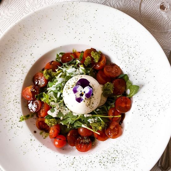 Ein weißer Teller mit einem Salat aus Tomaten, Basilikum und einer halbierten Burrata-Käse, gekrönt von einer violetten Blume, serviert auf einem weißen Tuch.