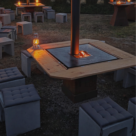 Außensitzbereich mit Holztischen und Stühlen, die um einen zentralen Feuerkorb mit einer Lampe herum angeordnet sind. Der Bereich ist von warmer Beleuchtung erhellt.