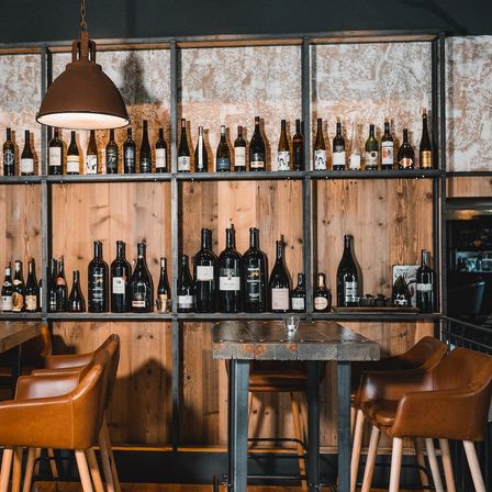 Eine Restaurantbar mit einer Wand aus Holzregalen, die viele Weinflaschen halten. Eine große Lampe hängt von der Decke. Es gibt Lederstühle und einen Tisch mit einem Glas darauf.