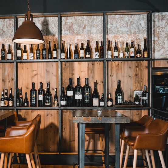 Eine Restaurantbar mit einer Wand aus Holzregalen, die viele Weinflaschen halten. Eine große Lampe hängt von der Decke. Es gibt Lederstühle und einen Tisch mit einem Glas darauf.
