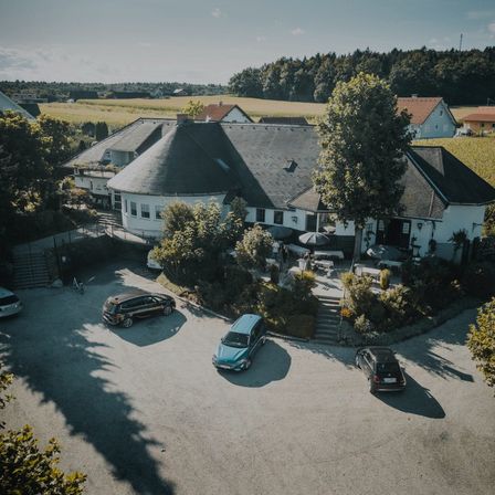 Ein Blick von oben auf ein Landhotel mit mehreren vor dem Gebäude geparkten Autos und einem Grasfeld im Hintergrund.