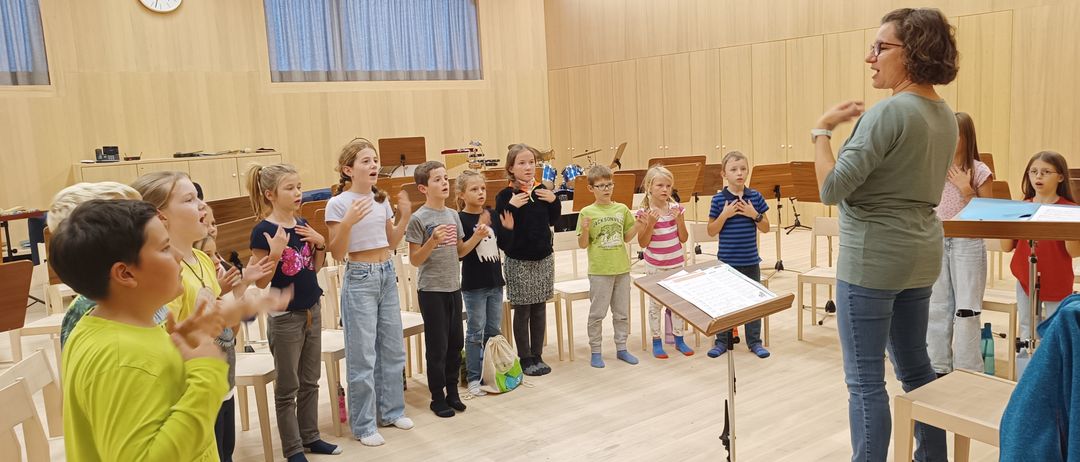 Mehrere Kinder stehen in einer Reihe und singen in einem Musikraum mit Holzwänden, Stühlen und Musikinstrumenten. Ein Notenständer mit einem Blatt Papier steht rechts.