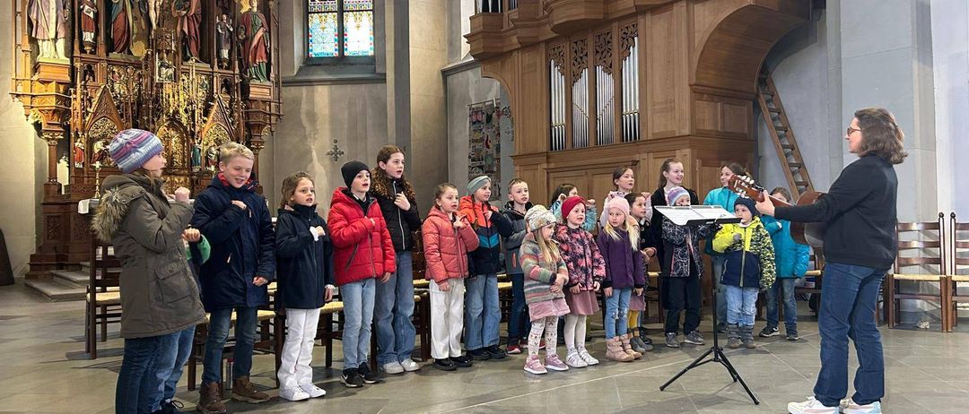 Ein Chor von Kindern in warmer Kleidung singt in einer Kirche. Eine Person spielt Gitarre vor ihnen.