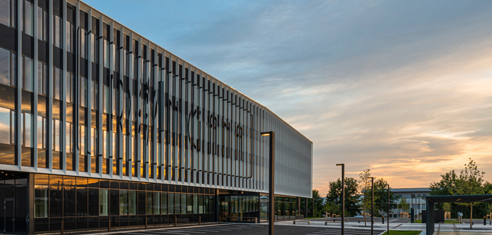 Ein modernes Bürogebäude mit einer Glasfassade, die den Sonnenuntergang widerspiegelt. Der davor liegende Parkplatz ist leer.