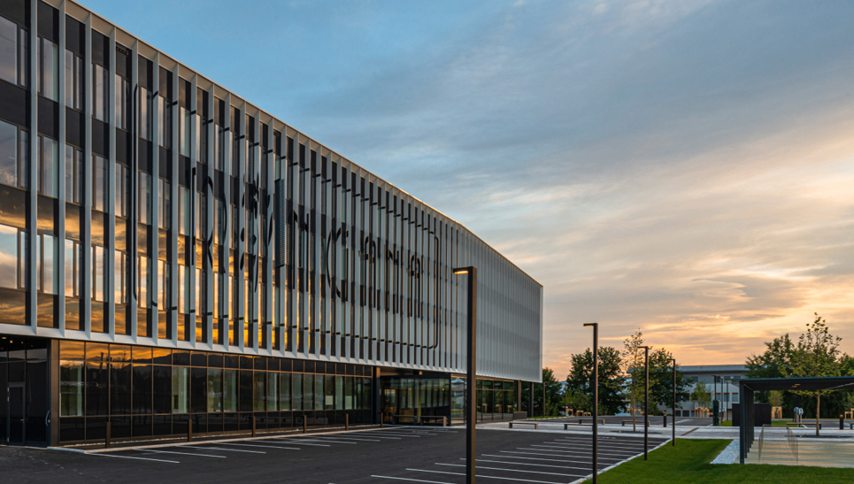 Ein modernes Bürogebäude mit einer Glasfassade, die den Sonnenuntergang widerspiegelt. Der davor liegende Parkplatz ist leer.