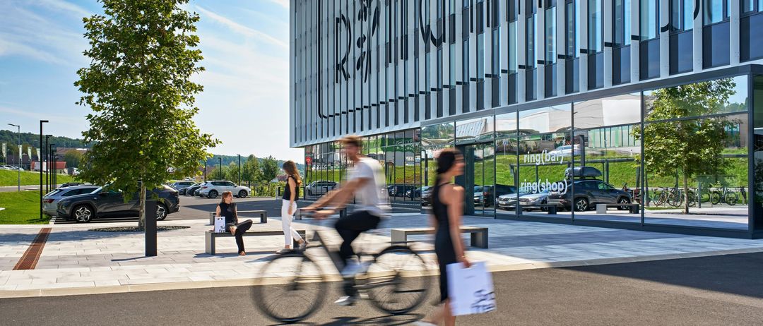 Ein Radfahrer fährt an einem modernen Gebäude vorbei, auf dem 'RSM' steht. Zwei Frauen gehen voraus, eine trägt einen Einkaufsbeutel. Bäume, geparkte Autos und Bänke sind ebenfalls vorhanden.