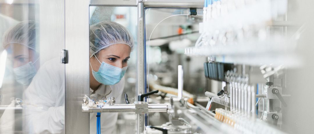 Eine Frau mit Maske und Haarnetz inspiziert eine Maschine in einer Fabrik und konzentriert sich auf ein Förderband mit kleinen Behältern.