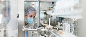 Eine Frau mit Maske und Haarnetz inspiziert eine Maschine in einer Fabrik und konzentriert sich auf ein Förderband mit kleinen Behältern.