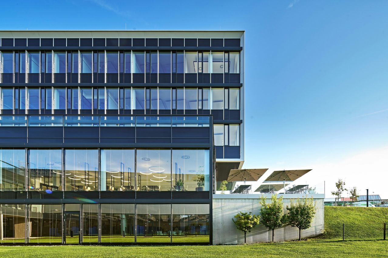 Ein modernes Bürogebäude mit Glasfassaden, die den blauen Himmel reflektieren. Es verfügt über mehrere Stockwerke, Außenbestuhlung mit Sonnenschirmen und einen Rasen.