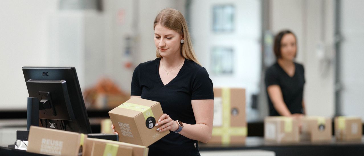 Eine Frau in einem schwarzen V-Ausschnitt-Shirt hält eine braune Box mit den Worten 'Reuse Concept' und 'Rethink'. Sie steht in einem Lagerhaus.