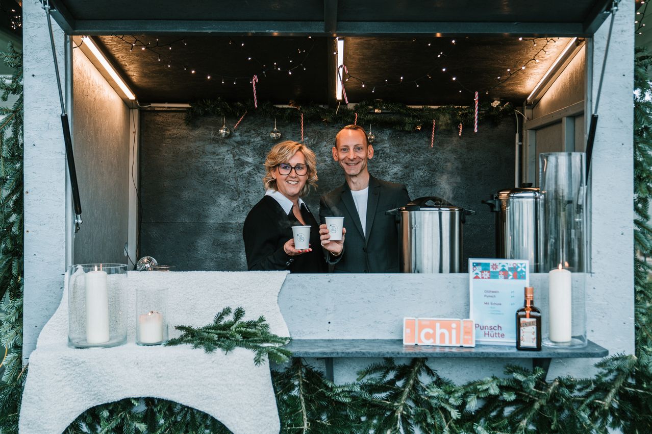 Ein Mann und eine Frau stehen lächelnd hinter einem festlich geschmückten Tresen mit Tassen. Dahinter stehen zwei dampfnudelnde Töpfe und ein Menüschild für 'Punsch Hutte'. Die Umgebung ist mit Weihnachtslichtern, Girlanden und einer Kerze dekoriert.