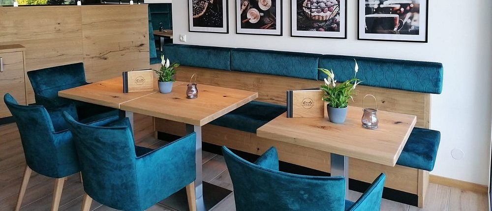 Ein gemütliches Restaurant mit Holztischen, blauen Samtstühlen und einer blauen Bank. Vier schwarze und goldene Pendelleuchten und vier gerahmte Bilder hängen an der Wand.
