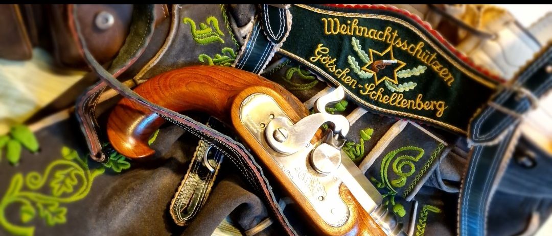 Eine Vintage-Pistole liegt auf einem traditionellen deutschen Kostüm mit aufwendiger Stickerei und einem festlichen Band mit der Aufschrift 'Weihnachtsschießen Goschen-Schellenber'.